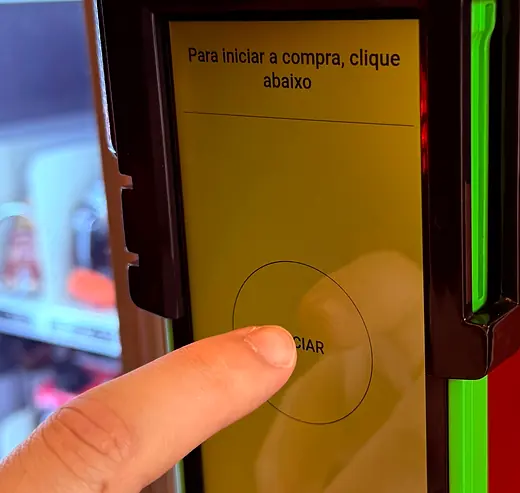 Dedo tocando a tela da máquina, que pede para clicar abaixo para iniciar a compra.